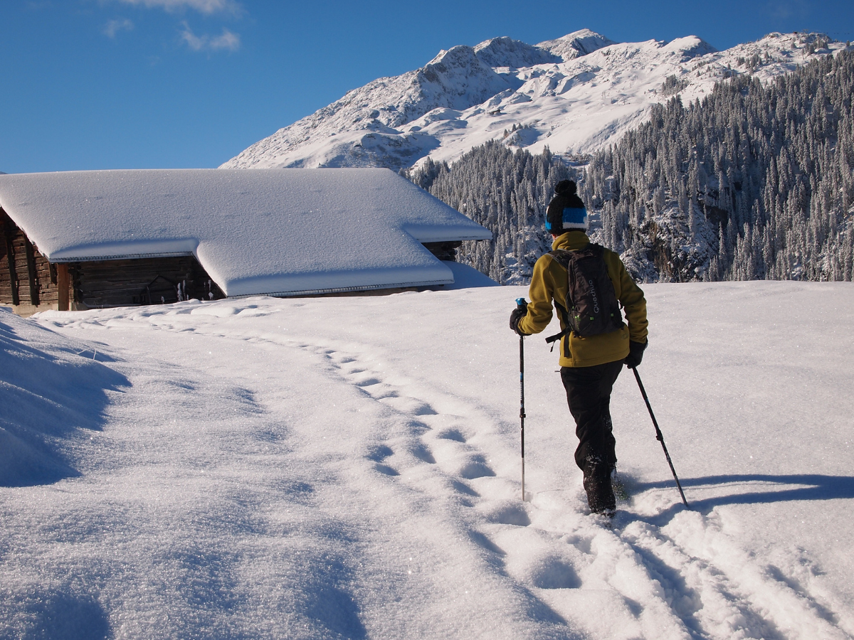 Un randonneur a raquettes suit un ancienne trace légèrement recouvert de neige fraiche. Il progresse en direction d'une grange à foin