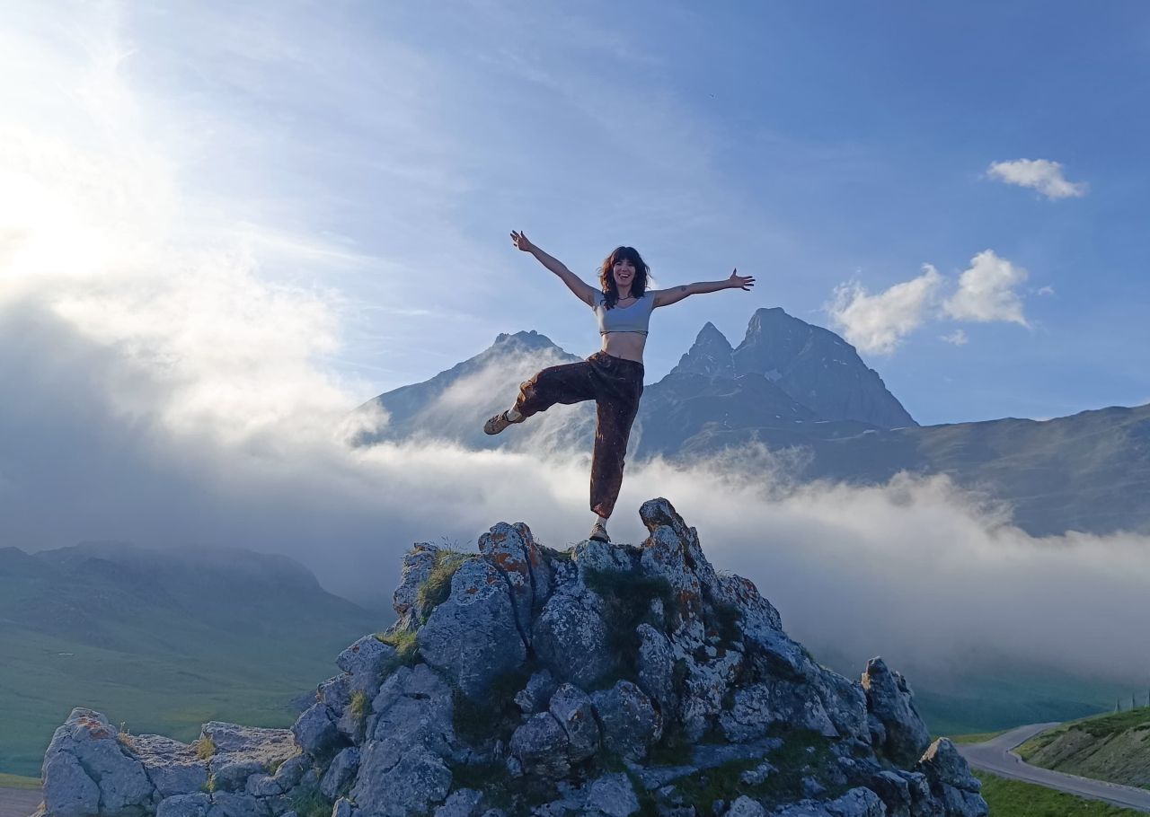 Jeune femme en équilibre sur un pied, bras écartés, au sommet d’un rocher face au Pic du Midi d’Ossau et à une mer de nuages